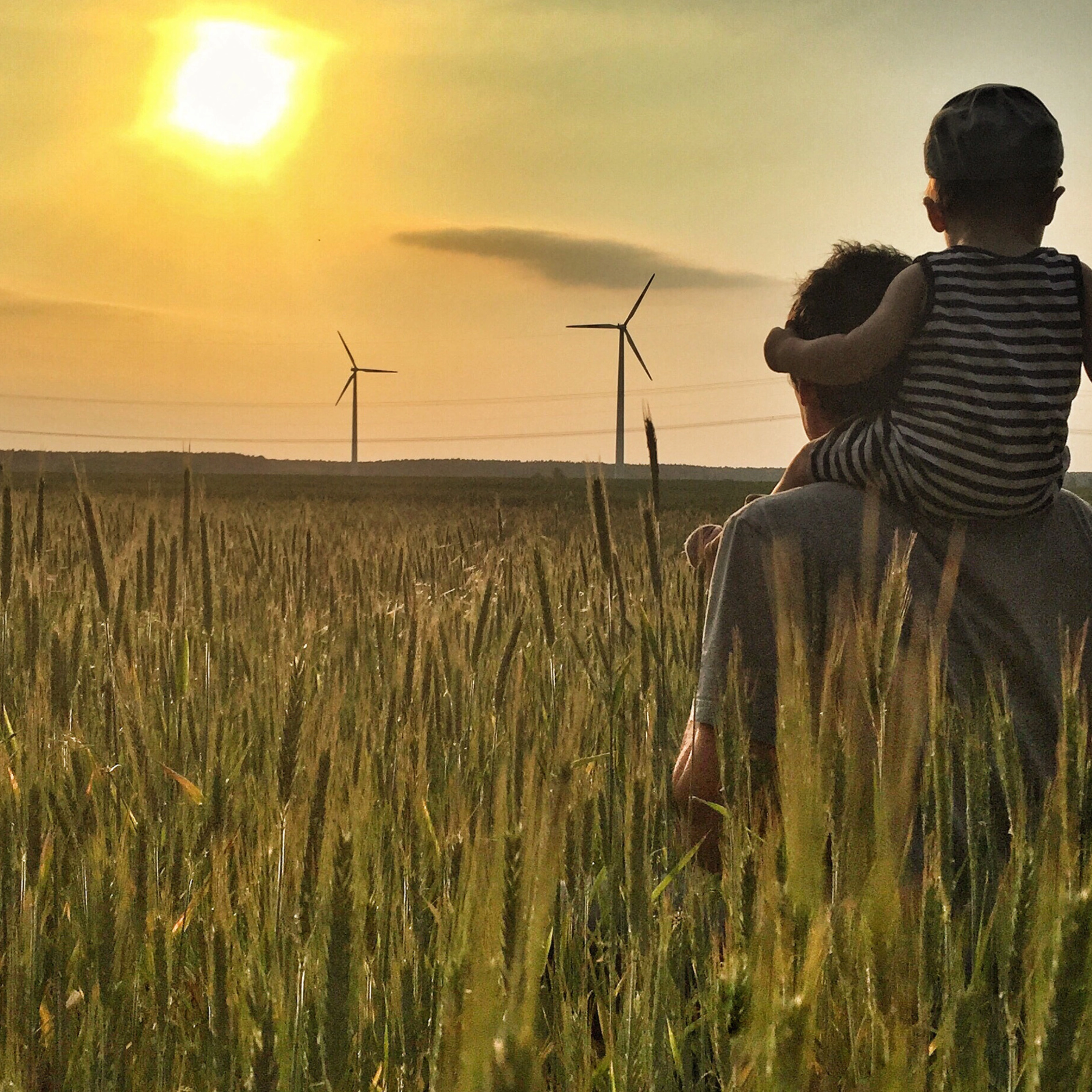 Man carries a child on his shoulders in a field at sunset with wind turbines in the background.