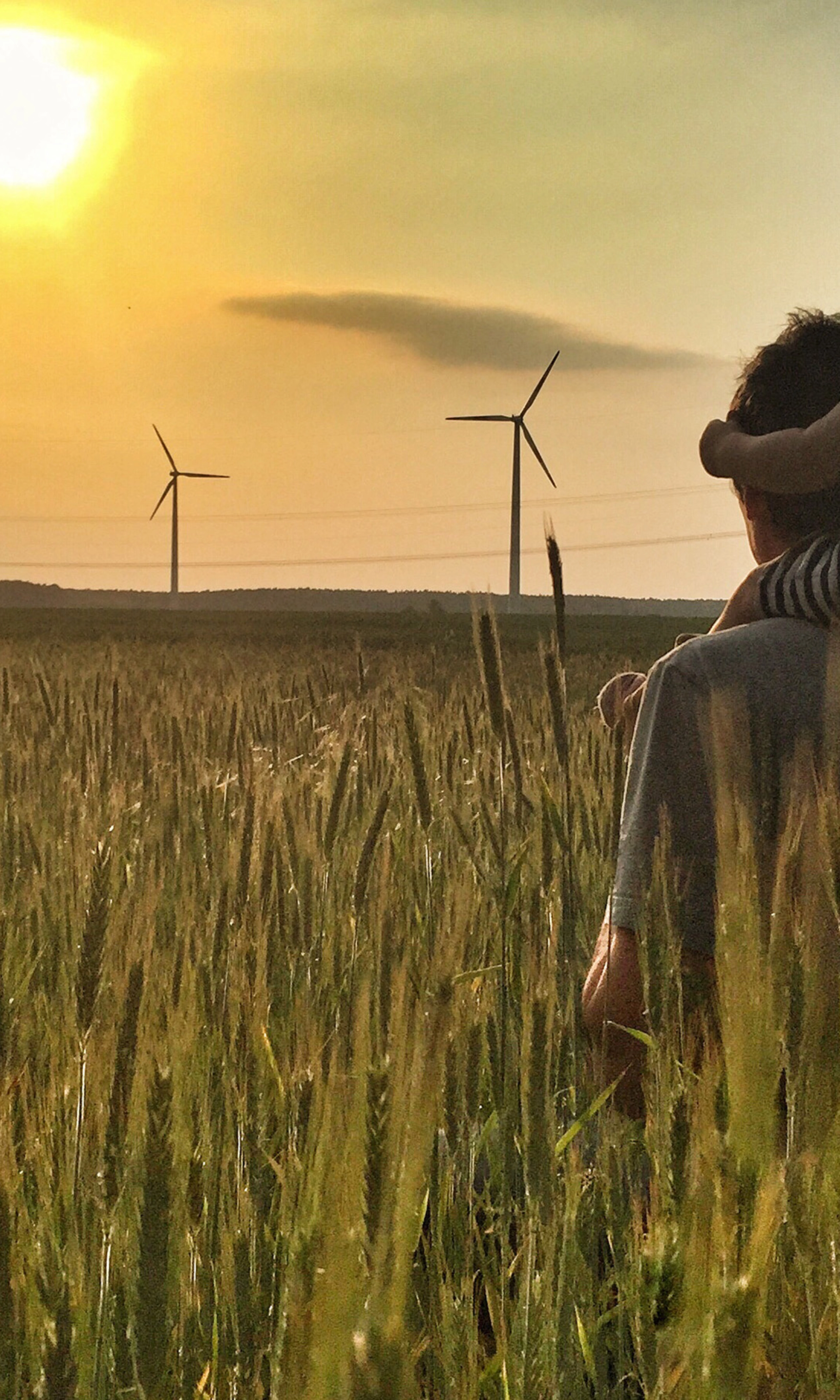 Man carries a child on his shoulders in a field at sunset with wind turbines in the background.