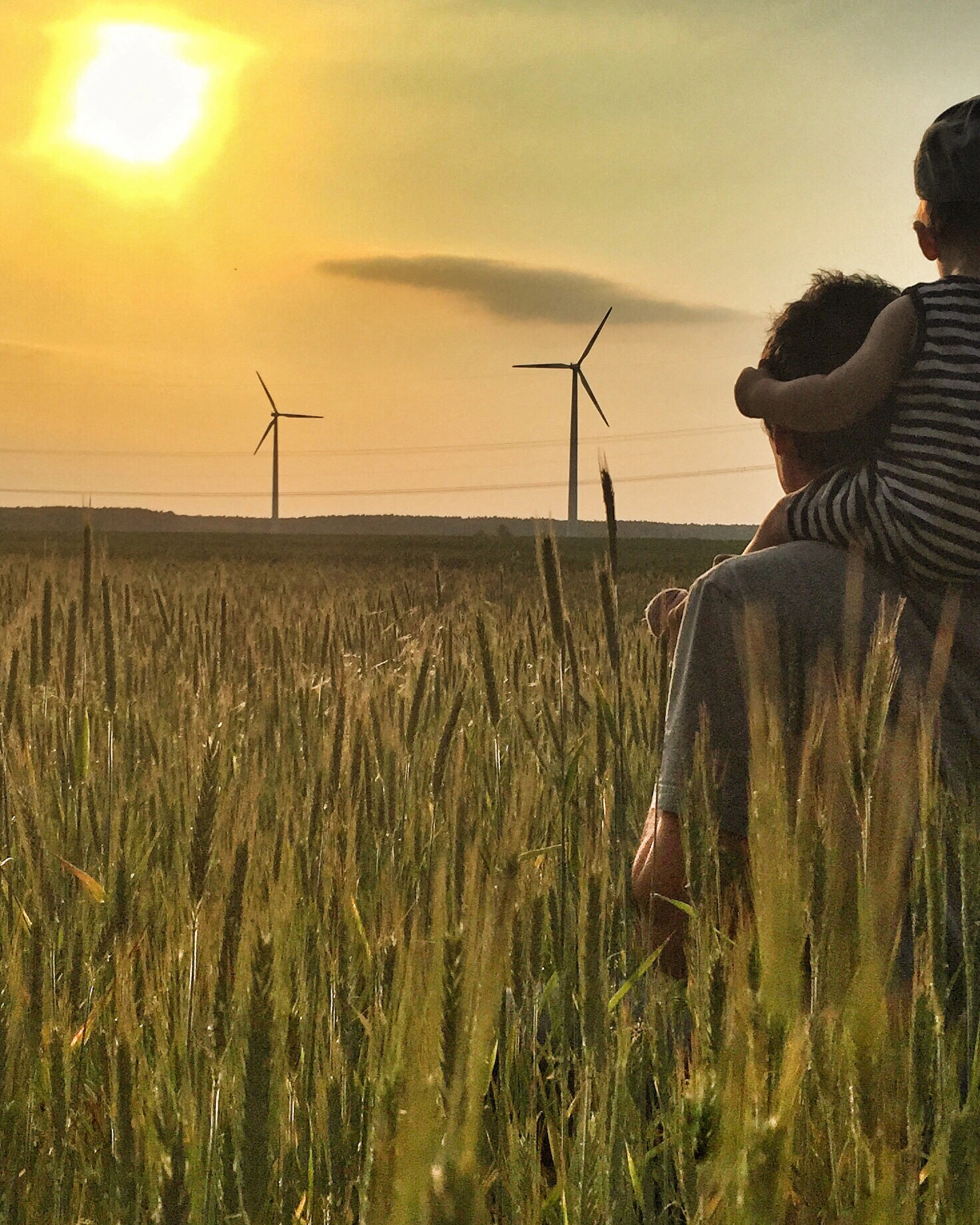 Man carries a child on his shoulders in a field at sunset with wind turbines in the background.