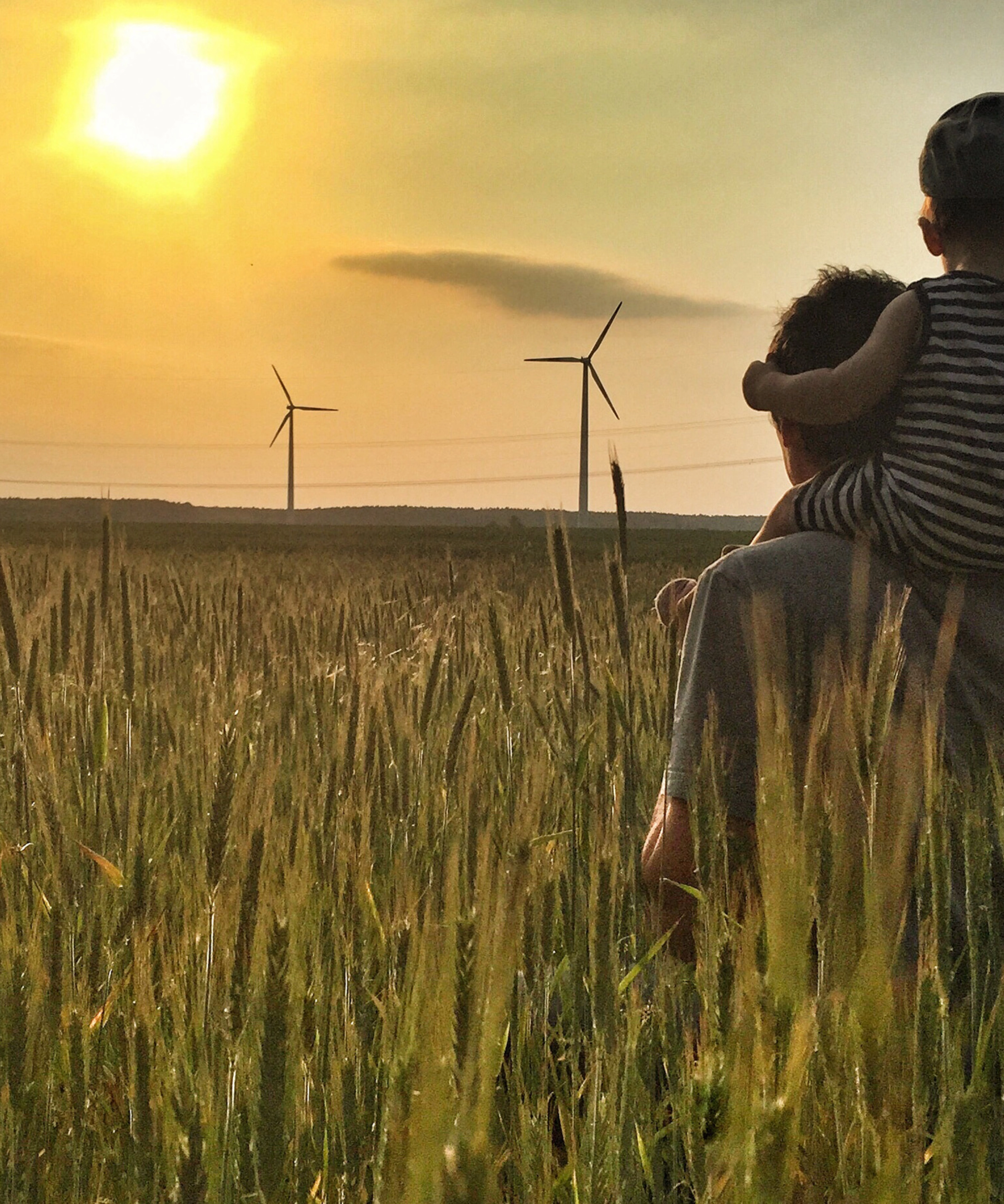 Man carries a child on his shoulders in a field at sunset with wind turbines in the background.