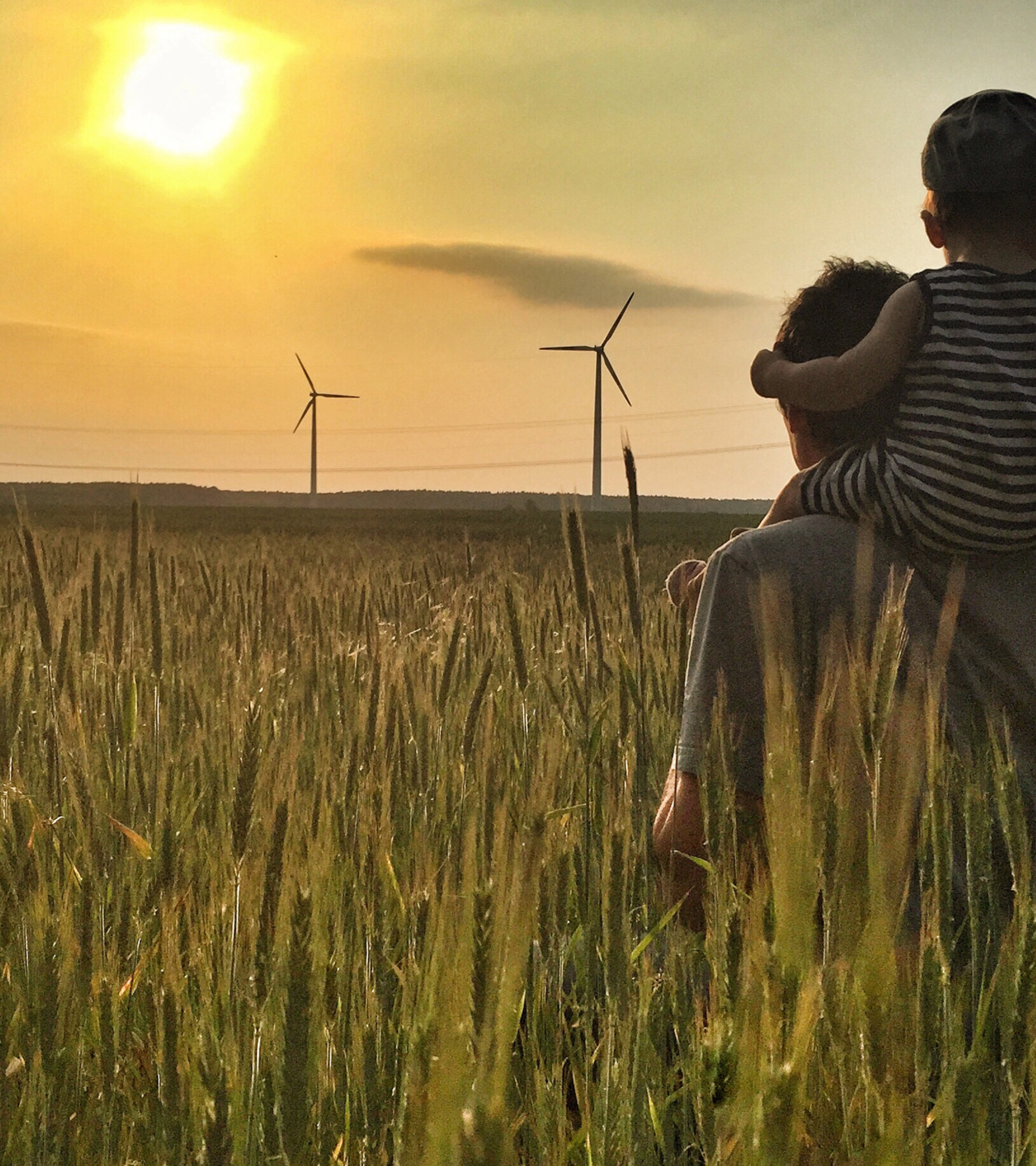Man carries a child on his shoulders in a field at sunset with wind turbines in the background.